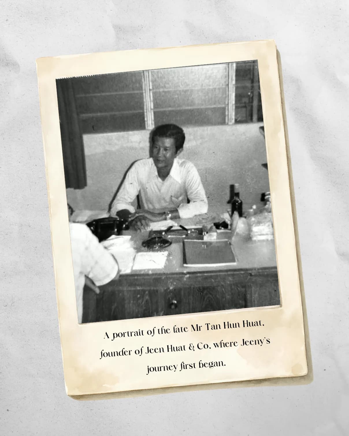 A black-and-white photograph of the late Mr Tan Hun Huat seated at his desk as a young man, printed on aged paper and captioned: a portrait of the late Mr Tan Hun Huat, founder of Jeen Huat & Co., where Jeeny's journey first began